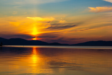 Obraz premium Golden sunset over tranquil lake with colorful sky and smooth reflections. Serene evening scene with mountain silhouettes, glowing horizon and peaceful nature. Perfect for travel and landscape themes.