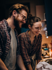 A smiling couple wearing glasses and plaid shirts enjoy a moment together in a cozy kitchen setting.