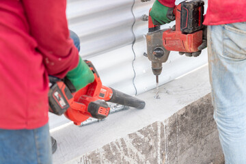 Two workers are using a cordless drill to drill into the concrete floor, while one of them is using a cordless blower to remove dust and debris.