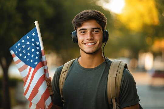 Smiling young man wearing headphones and a backpack holding a waving American flag outdoors in a sunny park environment