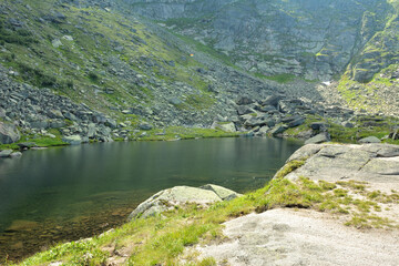 A small calm lake at the foot of a steep rocky slope of a high cliff on a sunny summer morning.