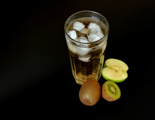 Tropical fruit juice in a tall glass with ice on a black background, next to it are pieces of ripe kiwi and green apple.