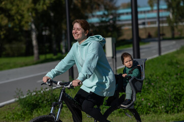 Caucasian woman riding bicycle with toddler son sitting behind her in child seat in park. 