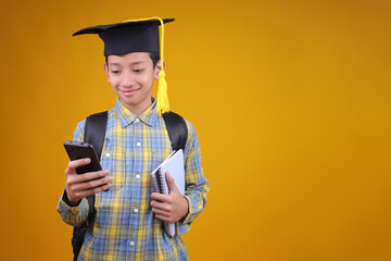 Smiling Young Asian Student In Graduation Cap Holding Notebooks While Using Mobile Phone
