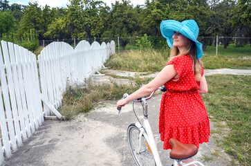 Young woman wearing a red dress and blue sun hat with a bicycle outdoors near a white fence is...