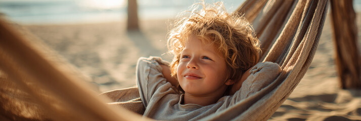 A young boy with curly hair relaxing in a hammock on a sandy beach near the ocean