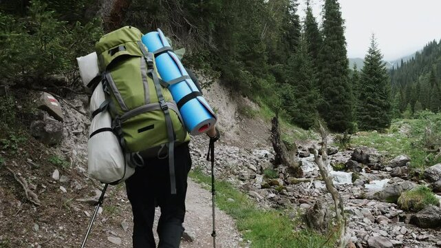 Backpacker hiking along mountain river trail with trekking poles and camping gear.