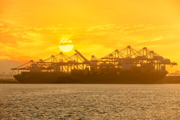 Cargo ships docked next to cranes with a sun setting in the background
