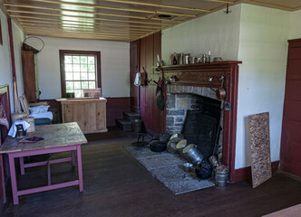 Interior of a traditional Canadian pioneer house with rustic wooden furniture, antique cookware, and a stone fireplace. A preserved historical home capturing 19th-century rural life.
