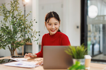 Smiling young asian woman sitting at desk working on laptop taking notes in notebook, happy...