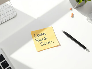 Yellow sticky note with Come Back Soon written on it is placed on white desk, surrounded by pen, keyboard, and plant