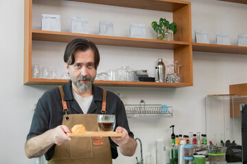 A confident senior coffee shop owner stands behind cafe bakery counter with arms crossed....