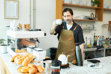 Senior Caucasian cafe owner in a brown apron raises his cup of coffee with a gentle smile. His welcoming presence and handcrafted setting reflect passion for personal service and small business pride