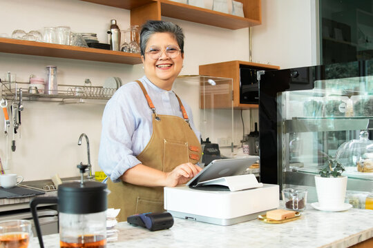 Asian senior woman using point-of-sale system behind the cafe counter with pastries nearby, representing modern technology in small food service and business management. Barista, business, technology.