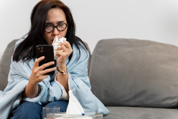 Sick woman blowing nose while using smartphone, wrapped in blanket on sofa, suffering from cold or...