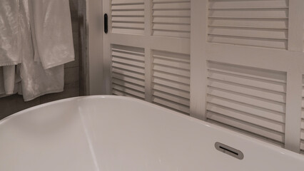 Bathroom interior. A fragment of a white oval acrylic bathtub. A partition made of blinds. Terry towels are suspended.  Shades of creamy beige. Close-up.