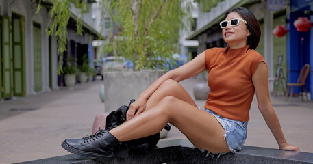Asian woman wearing sleeveless turtleneck top, distressed denim shorts and black combat boots enjoys the sun on a sleek stone bench in a charming urban alleyway