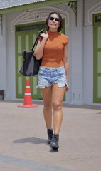 Fashionable Asian woman strides along a paved pathway in an urban environment, exuding confidence and style wear a fitted crop top paired with distressed denim shorts and combat boots