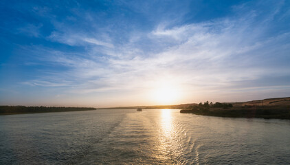 A beautiful sunset casts golden light over the tranquil Nile River near Esna Lock, Aswan, Egypt. A cruise boat is visible in the distance, sailing towards the lock at dusk.