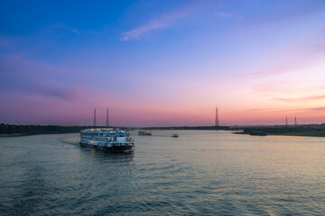 A large cruise ship glides along the tranquil Nile River near Esna Lock, Aswan, Egypt, at dusk. The sky above is painted with soft purple and pink hues, reflecting on the water.