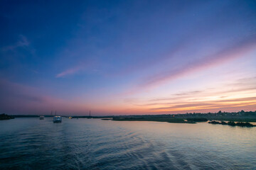 A tranquil wide shot of the Nile River at sunset near Esna Lock, Aswan, Egypt. The sky displays soft pink and purple hues, with silhouetted power towers and palm trees along the distant riverbanks.