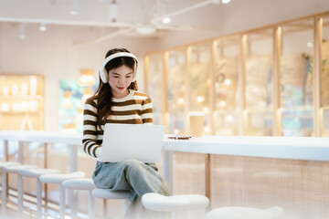 Young Asian woman sitting in a cozy café, smiling while using her smartphone and laptop, enjoying a hot drink, studying, working, and relaxing in a modern setting.