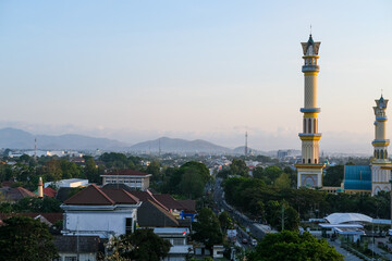 Obraz premium Evening light casts a golden hue over the Islamic Center Mosque in Mataram city, Lombok, with towering minarets, bustling streets, and distant hills completing the picturesque skyline