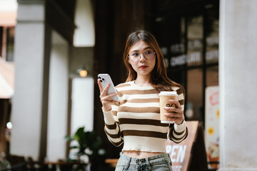 A stylish young woman, possibly an influencer, holds a coffee cup and uses her smartphone outdoors in an urban cafe environment.