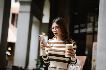 A stylish young woman, possibly an influencer, holds a coffee cup and uses her smartphone outdoors in an urban cafe environment.