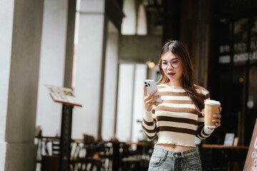 A stylish young woman, possibly an influencer, holds a coffee cup and uses her smartphone outdoors in an urban cafe environment.