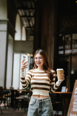 A stylish young woman, possibly an influencer, holds a coffee cup and uses her smartphone outdoors in an urban cafe environment.