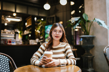 A stylish young woman with glasses and braces smiles at the camera, holding a coffee cup in a warmly lit cafe setting.