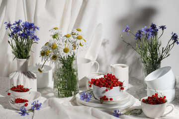 Bouquets of cornflowers and daisies in vases on the table and red currants in white bowls. Summer still life.