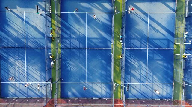 Aerial view of three vibrant blue padel courts, captured by a drone. Several players are actively engaged in padel matches, moving energetically across the courts. The scene is framed with strong