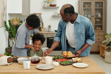 Two children with their father setting breakfast table with various foods, including croissants and fruit, in spacious modern kitchen. Family bonding in warm home environment