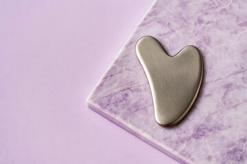Fototapeta premium Elegant stainless steel gua sha facial massage tool displayed on a textured purple marble surface with a soft lavender backdrop, representing luxury skincare, beauty rituals, and holistic wellness.