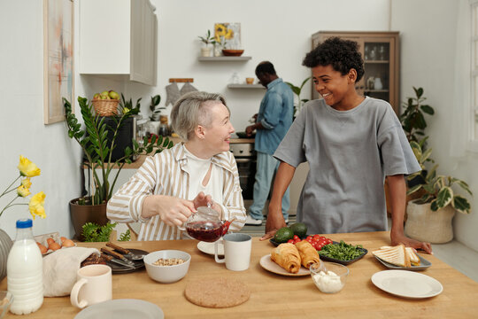 Woman pouring drink while chatting with smiling child in kitchen setting with another person cooking in background seen showcasing family bonding and happiness
