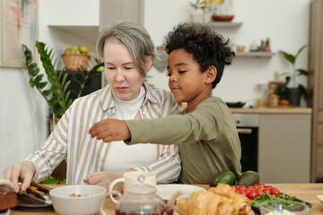 Caucasian woman with senior characteristics and African American child bonding over preparing...