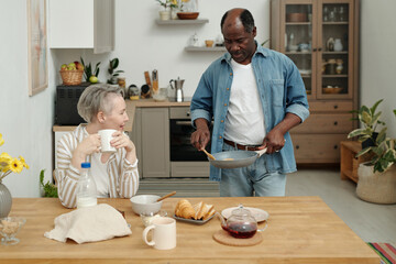 Mature woman enjoying coffee while standing man serving breakfast at home. Modern kitchen with wooden elements and various kitchenware present