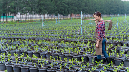 Asian female farmer walking through potted plant nursery, carrying hoe on shoulder, smiling after...