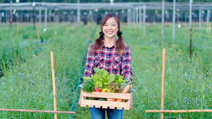 Happy Asian female farmer holding wooden crate full of fresh organic vegetables, smiling in green farmland, sustainable agriculture and healthy food concept