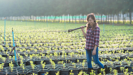 Young female farmer walking through plant nursery with pitchfork over shoulder, symbolizing hard work, sustainable agriculture, and eco-friendly farming lifestyle