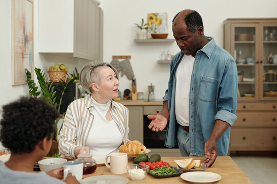 Middle-aged couple and their son having breakfast at kitchen table discussing plans for day. Fresh and healthy ingredients like avocado, berries, and bread laid out