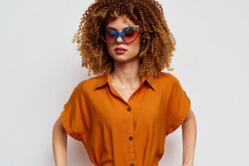 Fashionable woman with curly hair wearing stylish sunglasses and a stylish orange shirt, posing...
