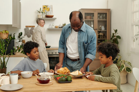 Diverse family sharing breakfast in cozy kitchen, with elder preparing food and children engaged in conversation. Scene captures warm family dynamics and cheerful atmosphere