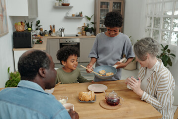 Multiethnic family sharing breakfast at home, with young boy serving pancakes. Smiling and engaging with breakfast items on rustic wooden table