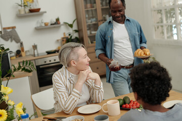 Caucasian woman enjoying meal with family while African American man serving beverage, sitting at kitchen table filled with food, flowers, and kitchenware