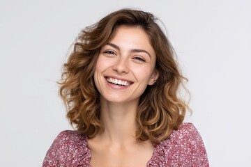 A woman smiles widely revealing her teeth in a closeup studio shot against a plain backdrop She has wavy brown hair and is wearing a floral blouse