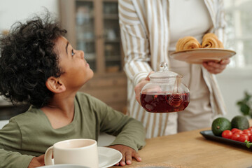Young child smiling while sitting at kitchen table with breakfast, looking up at parent holding tea...