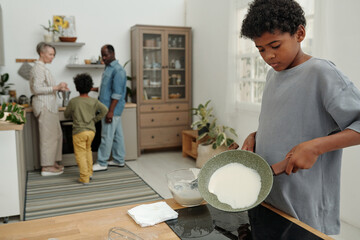 Family engaged in cooking activity in modern kitchen with child pouring ingredients into pan while...